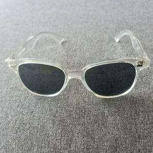 Peut inclure: Lunettes de soleil &agrave; monture transparente avec verres fonc&eacute;s. Les lunettes ont un design classique et r&eacute;tro avec une monture et des branches l&eacute;g&egrave;rement incurv&eacute;es. Les verres sont noirs, contrastant avec la monture transparente. Un accessoire &eacute;l&eacute;gant.