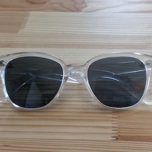 Peut inclure: Lunettes de soleil &agrave; monture transparente avec verres fonc&eacute;s. Les lunettes ont une forme ronde classique et sont en plastique transparent. Les verres semblent teint&eacute;s. Les lunettes reposent sur une surface en bois.