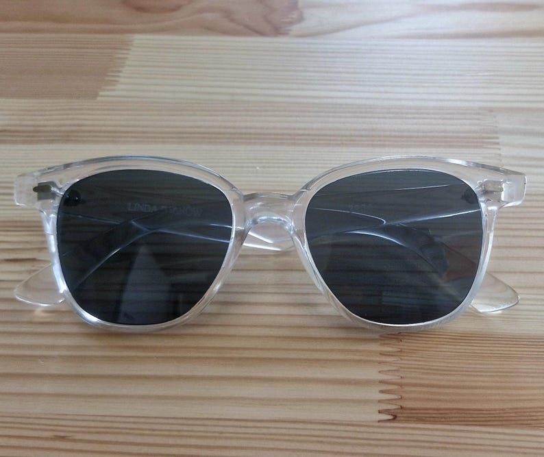 Peut inclure: Lunettes de soleil &agrave; monture transparente avec verres fonc&eacute;s. Les lunettes ont une forme ronde classique et sont en plastique transparent. Les verres semblent teint&eacute;s. Les lunettes reposent sur une surface en bois.