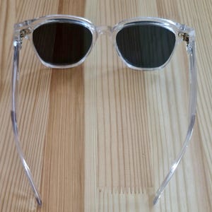 Peut inclure: Lunettes de soleil &agrave; monture transparente avec verres fonc&eacute;s. Les lunettes ont une forme carr&eacute;e classique, l&eacute;g&egrave;rement arrondie. Les branches sont fines et transparentes, et les charni&egrave;res sont dor&eacute;es. Les lunettes reposent sur une surface en bois clair.
