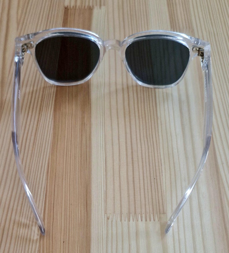 Peut inclure: Lunettes de soleil &agrave; monture transparente avec verres fonc&eacute;s. Les lunettes ont une forme carr&eacute;e classique, l&eacute;g&egrave;rement arrondie. Les branches sont fines et transparentes, et les charni&egrave;res sont dor&eacute;es. Les lunettes reposent sur une surface en bois clair.