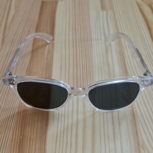 Peut inclure: Lunettes de soleil &agrave; monture transparente avec verres fonc&eacute;s. Les lunettes ont une forme classique, l&eacute;g&egrave;rement &oelig;il de chat. Les montures et les branches sont transparentes, et les verres sont d'une couleur fonc&eacute;e et unie. Les lunettes reposent sur une surface en bois clair.