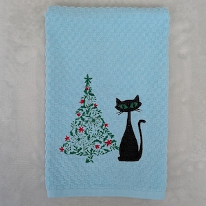 May include: A light blue waffle-weave kitchen towel featuring an embroidered design of a stylized green Christmas tree with red floral accents and a black cat with bright green eyes sitting upright next to the tree on the fabric surface.