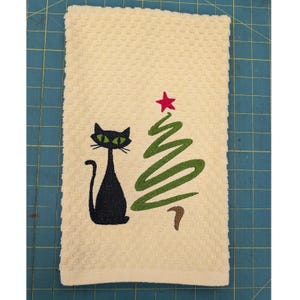 May include: A cream-colored waffle-weave kitchen towel featuring an embroidered black cat with bright green eyes positioned next to a stylized green Christmas tree topped with a small red star. The design is centered on the rectangular fabric.