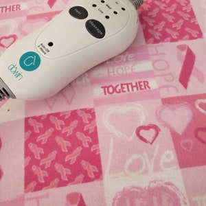May include: White electric heating pad controller with a blue logo that says "dowin". The controller has buttons for heat, massage, and an auto-off timer. The controller is on a pink and white fabric with a pattern of hearts and pink ribbons.