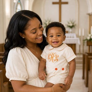 May include: A smiling person holds a baby wearing a cream-colored romper with orange embroidery and buttons. The baby is also wearing beige socks and brown shoes. The background shows a church interior with a cross and pews.