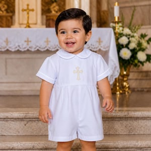 May include: A young child in a white romper suit, featuring a cross emblem on the chest. The outfit has a Peter Pan collar and short sleeves. The child is wearing white socks and white leather shoes. The background suggests a church interior.