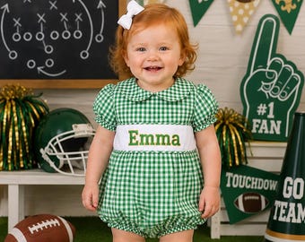 May include: A green and white gingham bubble romper with a white chest panel featuring the embroidered name Emma in green letters. The outfit includes a white hair bow, white lace-trimmed socks, and white shoes, creating a coordinated look.