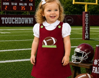May include: A maroon jumper dress featuring a white collared shirt underneath, green buttons at the shoulders, and an embroidered football graphic on the front. The outfit is paired with white tights and white shoes, with a white hair bow.