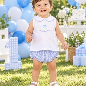 May include: A white sleeveless shirt with a Peter Pan collar featuring blue gingham trim and an embroidered blue cake with the text Birthday Boy, paired with blue and white gingham bloomers and white sandals for a coordinated outfit.