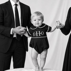 May include: A black romper with white trim and the letters "E.H.O" on the front. The baby is standing on a white surface, held by two adults in black suits. The image is in black and white.