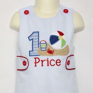May include: A blue and white striped romper with a red button closure. The romper has an embroidered design of a beach scene with a beach ball, bucket, and crab. The number "1" is also embroidered on the romper. The name "Price" is embroidered below the beach scene.