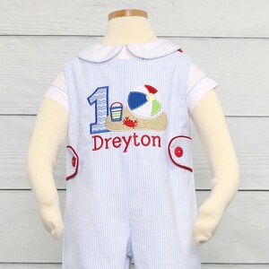 May include: A blue and white striped romper with a white collar and short sleeves. The romper has a red and white embroidered number one with a beach scene and the name "Dreyton" embroidered below. The romper is on a white mannequin.