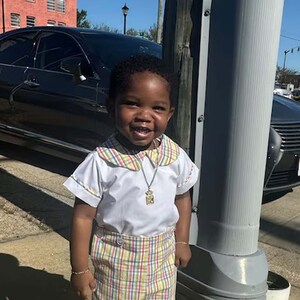 May include: A young child wearing a white shirt with a Peter Pan collar and plaid shorts. The outfit is completed with white socks, cream-colored shoes, and a gold pendant necklace. The child is smiling and standing outside on a sunny day.