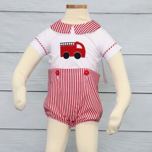 May include: A white short-sleeved shirt with a red and white striped collar and red and white striped cuffs. The shirt features a red fire truck applique on the front. The shirt is worn with red and white striped shorts with red buttons. The outfit is displayed on a mannequin with white socks and white shoes.