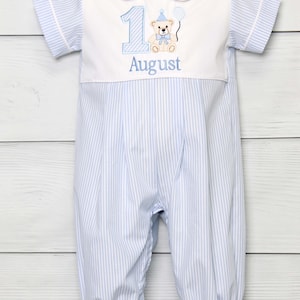 May include: Blue and white striped romper with a Peter Pan collar. The romper features a white panel with the number 1, a teddy bear, and the word "August" embroidered in blue. The outfit has short sleeves and gathered cuffs.