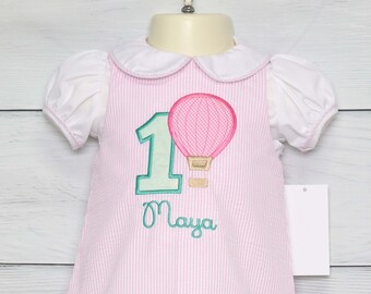 May include: Pink and white striped dress with a green number one and a hot air balloon design. The name "Maya" is embroidered below the number one.
