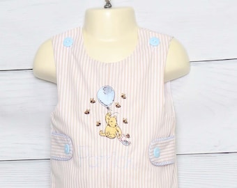 Puede incluir: Romper de bebé a rayas rosas y blancas, con un diseño de Winnie the Pooh. El romper presenta un dibujo de Winnie the Pooh sosteniendo un globo azul, rodeado de abejas. El nombre "Patrick" está bordado en la parte delantera. Botones y ribetes azul claro.
