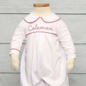 May include: White baby romper with long sleeves and a rounded collar. The collar and cuffs are trimmed with red gingham. The name "Coleman" is embroidered in red script across the chest. The romper has a gathered waist and elasticized ankles.