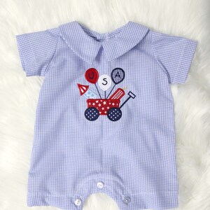 May include: A baby romper in a light blue and white gingham pattern. The romper features a Peter Pan collar and short sleeves. An embroidered design of a red wagon with fireworks and balloons spelling "USA" is on the front.
