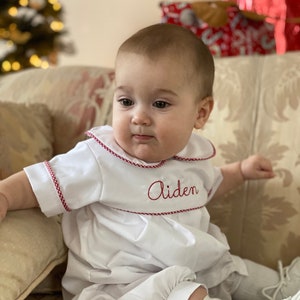 May include: A white baby romper with red trim and the name "Aiden" embroidered in red. The romper has a Peter Pan collar and short sleeves. The baby is wearing white socks and shoes.