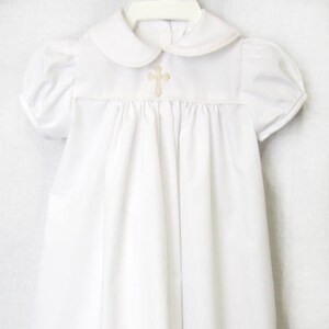 May include: White baptism gown featuring a Peter Pan collar and short, puffed sleeves. A gold-coloured cross is embroidered on the bodice. The dress is made of a soft fabric and hangs on a white hanger.