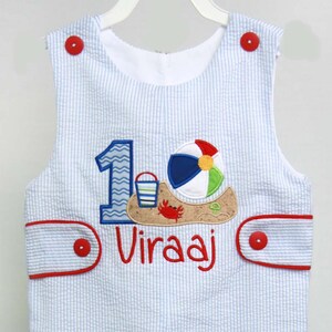 May include: Blue and white striped overalls with a red trim and buttons. The overalls have an embroidered number "1" with a beach scene and the name "Viraaj" on the front.