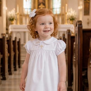 May include: A young child wearing a white dress with a Peter Pan collar and a gold cross emblem. The dress has short sleeves and a pleated skirt. The child is wearing white shoes and socks with frills. A white bow is in the child's hair.
