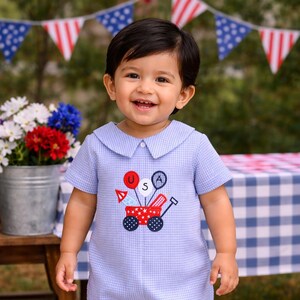 May include: A smiling toddler wearing a light blue gingham romper with a patriotic wagon design. The romper features a Peter Pan collar and short sleeves. The child is wearing navy sandals and is standing outdoors.