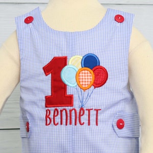 May include: A blue and white gingham baby onesie with a red number one and the name "Bennett" embroidered on the front. The onesie has red buttons on the shoulders and is decorated with colorful balloons.