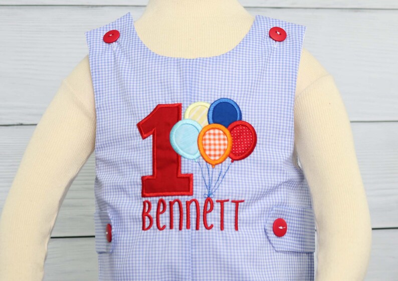 May include: A blue and white gingham baby onesie with a red number one and the name "Bennett" embroidered on the front. The onesie has red buttons on the shoulders and is decorated with colorful balloons.