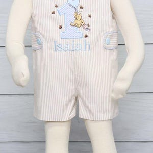 May include: A light brown and white striped baby romper with blue trim and buttons. The romper features an embroidered Winnie the Pooh character holding a balloon and the number one. The romper is personalized with the name "Isaiah".