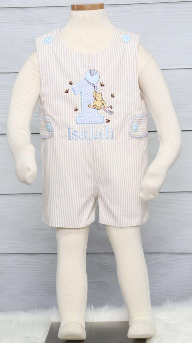 May include: A light brown and white striped baby romper with blue trim and buttons. The romper features an embroidered Winnie the Pooh character holding a balloon and the number one. The romper is personalized with the name "Isaiah".