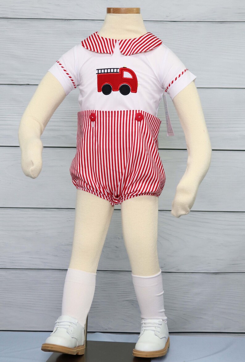 May include: A white short-sleeved shirt with a red and white striped collar and red and white striped cuffs. The shirt features a red fire truck applique on the front. The shirt is worn with red and white striped shorts with red buttons. The outfit is displayed on a mannequin with white socks and white shoes.