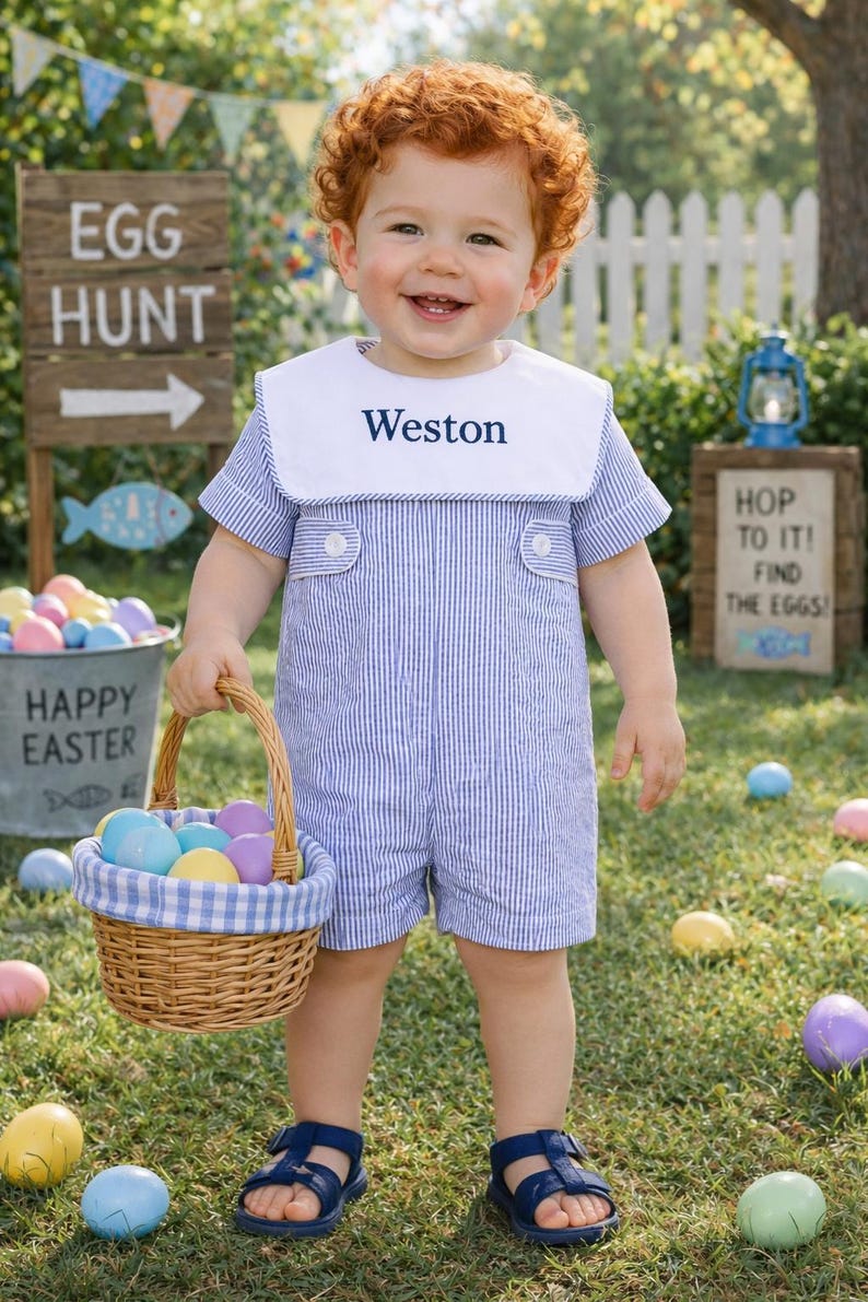 May include: A young child wearing a blue and white striped romper with a white collar, holding a basket of Easter eggs. The romper has the name "Weston" embroidered on the collar. The child is wearing blue sandals and is standing in a grassy area with Easter decorations.