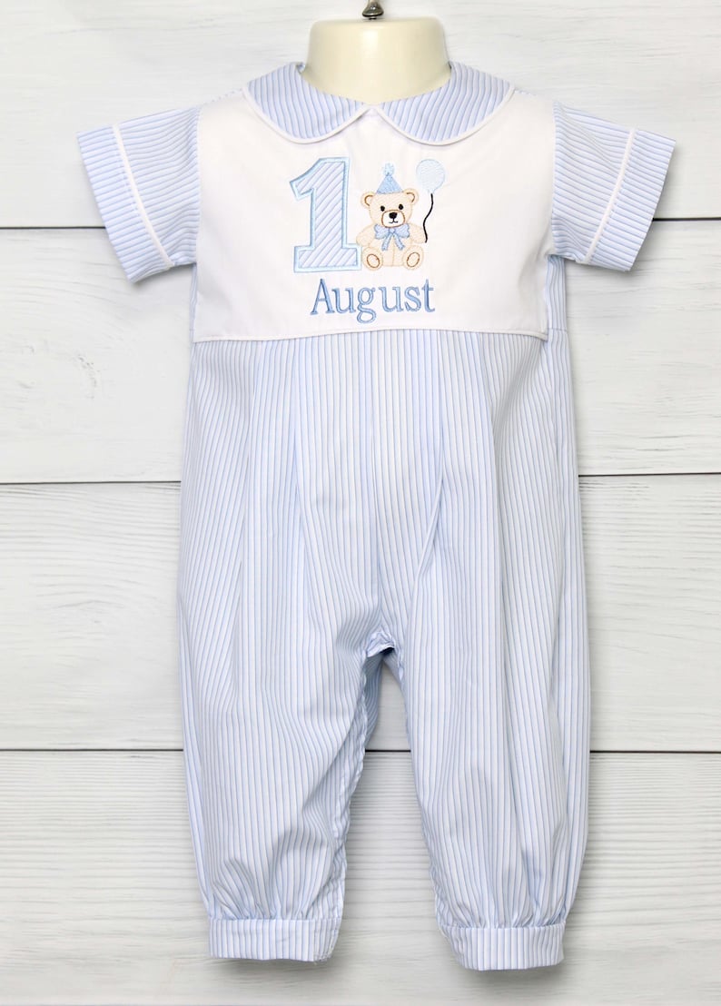 May include: Blue and white striped romper with a Peter Pan collar. The romper features a white panel with the number 1, a teddy bear, and the word "August" embroidered in blue. The outfit has short sleeves and gathered cuffs.