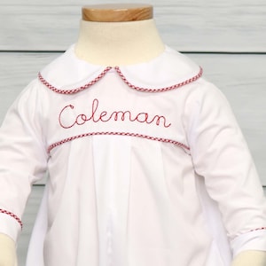 May include: White baby romper with long sleeves, a Peter Pan collar, and red trim. The name "Coleman" is embroidered in red script across the chest. The outfit is displayed on a mannequin.