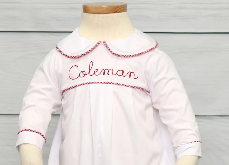 May include: White baby romper with long sleeves, a Peter Pan collar, and red trim. The name "Coleman" is embroidered in red script across the chest. The outfit is displayed on a mannequin.