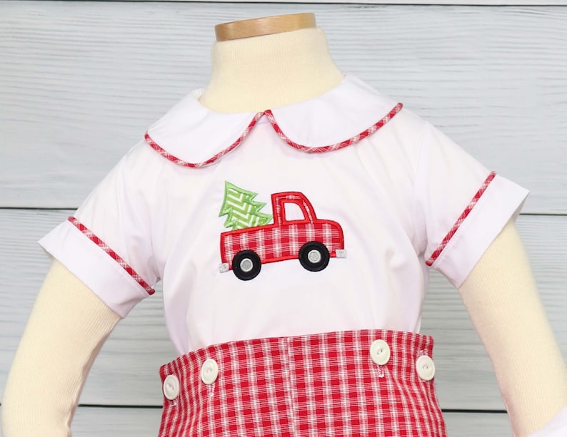 Puede incluir: Conjunto de camisa blanca y pantal&oacute;n corto a cuadros rojos. La camisa presenta un cami&oacute;n rojo con un &aacute;rbol de Navidad verde. El cuello y las mangas est&aacute;n ribeteados con cuadros rojos. El pantal&oacute;n corto tiene botones blancos.