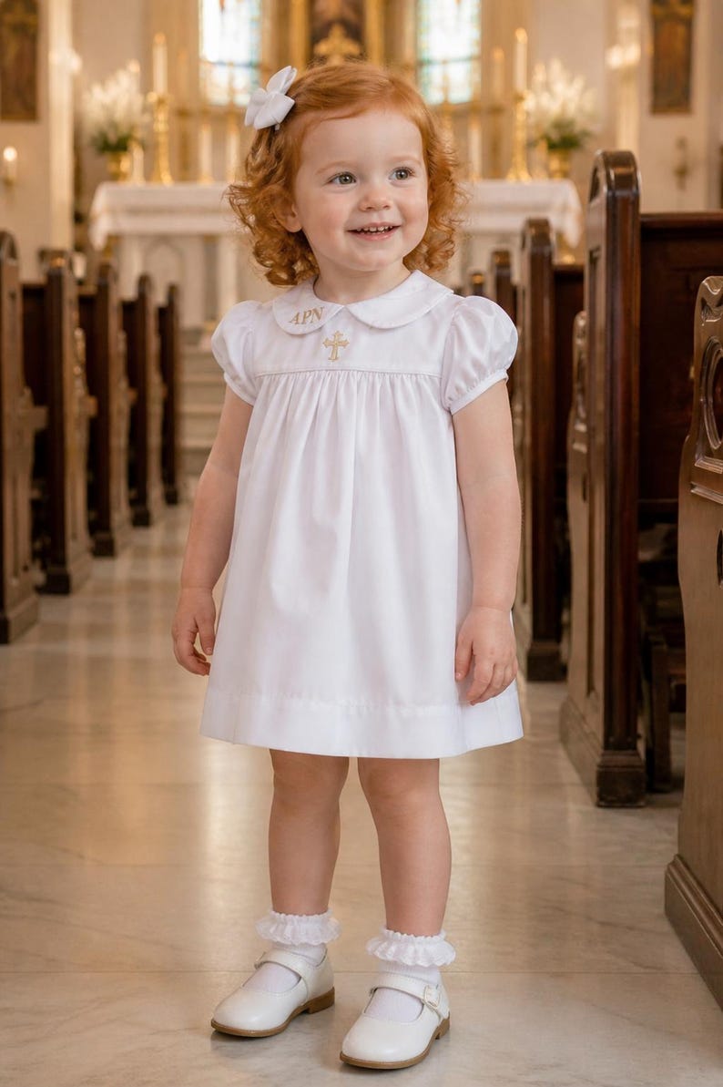 May include: A young child wearing a white dress with a Peter Pan collar and a gold cross emblem. The dress has short sleeves and a pleated skirt. The child is wearing white shoes and socks with frills. A white bow is in the child's hair.