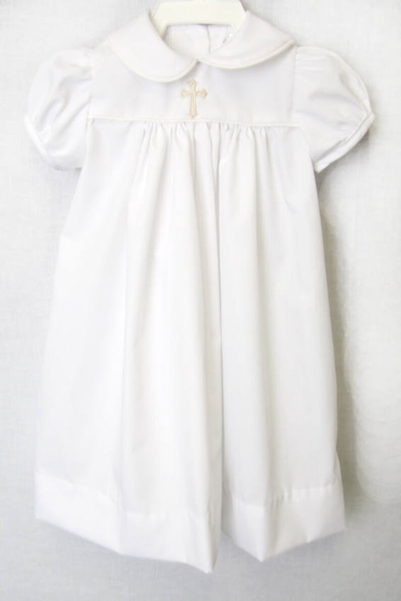 May include: White baptism gown featuring a Peter Pan collar and short, puffed sleeves. A gold-coloured cross is embroidered on the bodice. The dress is made of a soft fabric and hangs on a white hanger.