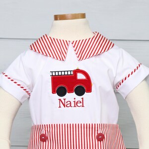 May include: A white short-sleeved outfit with red and white striped trim. The outfit has a red fire truck embroidered on the front with the name "Naiel" embroidered below it.