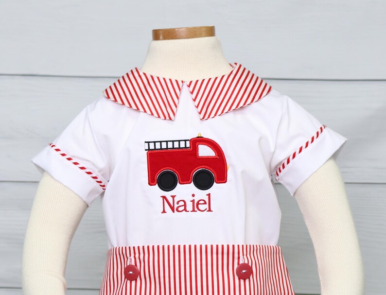 May include: A white short-sleeved outfit with red and white striped trim. The outfit has a red fire truck embroidered on the front with the name "Naiel" embroidered below it.