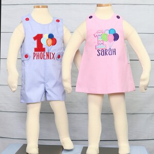 May include: Two baby outfits for a first birthday. A blue and white gingham romper with the number one and the name "Phoenix" embroidered on it. A pink gingham dress with the number one and the name "Sarah" embroidered on it. Both outfits feature colorful balloons.