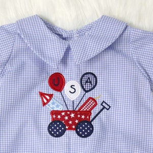 May include: Blue and white gingham romper with a patriotic design. The embroidered design features a red wagon with fireworks and balloons spelling "USA". The romper has a Peter Pan collar and short sleeves.