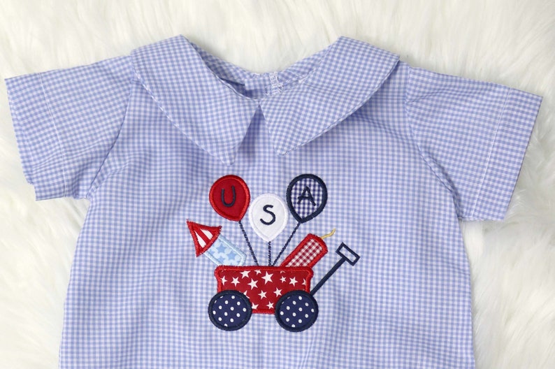 May include: Blue and white gingham romper with a patriotic design. The embroidered design features a red wagon with fireworks and balloons spelling "USA". The romper has a Peter Pan collar and short sleeves.