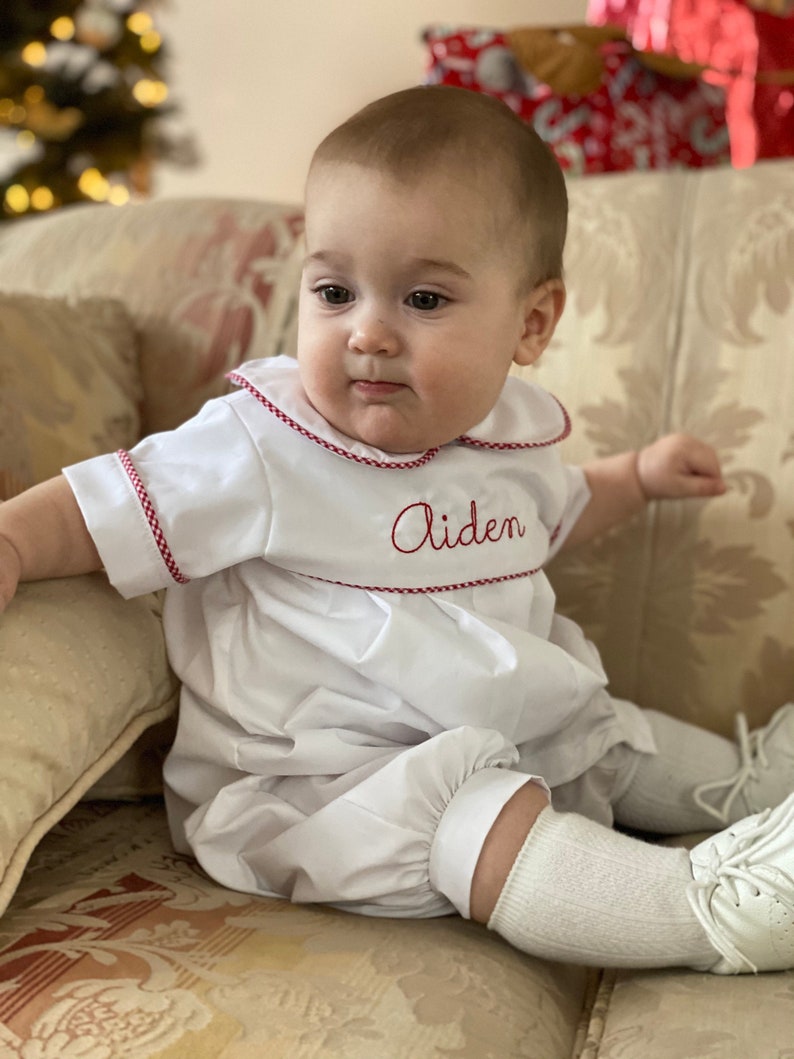 May include: A white baby romper with red trim and the name "Aiden" embroidered in red. The romper has a Peter Pan collar and short sleeves. The baby is wearing white socks and shoes.