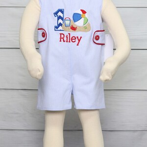 May include: A blue and white striped romper with red trim and a red button closure. The romper has an embroidered design of a beach ball, a bucket and shovel, and a crab. The number "1" is also embroidered on the romper, along with the name "Riley".