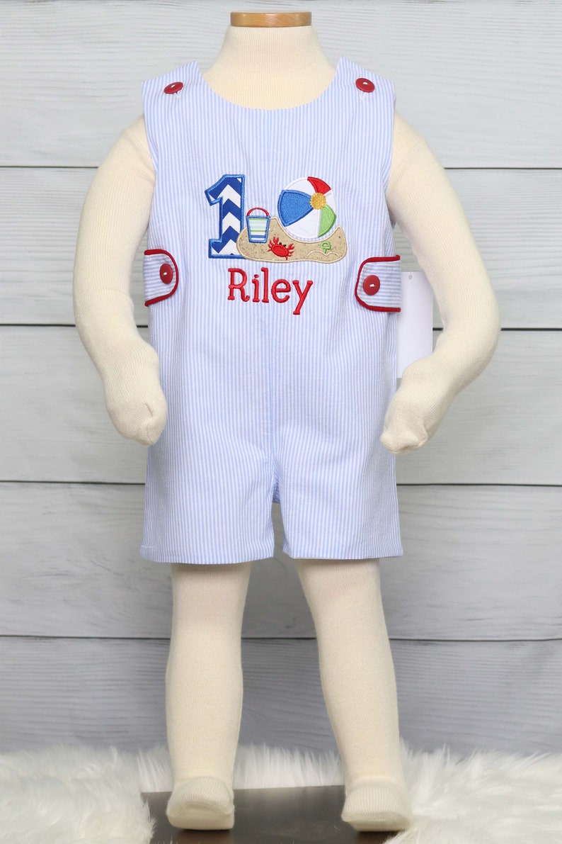 May include: A blue and white striped romper with red trim and a red button closure. The romper has an embroidered design of a beach ball, a bucket and shovel, and a crab. The number "1" is also embroidered on the romper, along with the name "Riley".