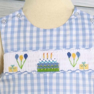 May include: A blue and white gingham baby onesie with a white embroidered design of a birthday cake with candles, balloons, and a gift box.